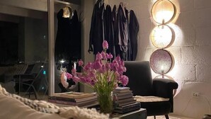Una camera accogliente con un'ampia finestra, una poltrona nera, un comodino con libri e un vaso di fiori, e un lampadario a parete.
