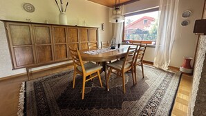 Un coin repas avec une table en bois, des chaises et un grand tapis. Il y a une fenêtre qui donne sur une maison et un toit rouge.