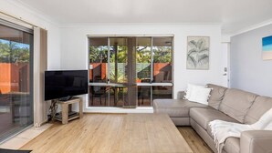 Una sala de estar moderna con un televisor de pantalla plana, un sofá color beige, una mesa de centro de madera y un patio exterior con una mesa y sillas al que se accede por una puerta corrediza de vidrio.