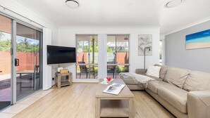 Una sala de estar moderna con un televisor de pantalla plana, un sofá color beige, una mesa de centro de madera y un gran ventanal corredizo que da a un espacio exterior.