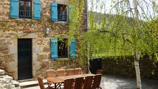 Un edificio in pietra con persiane blu, un tavolo di legno con sedie e un albero sullo sfondo.