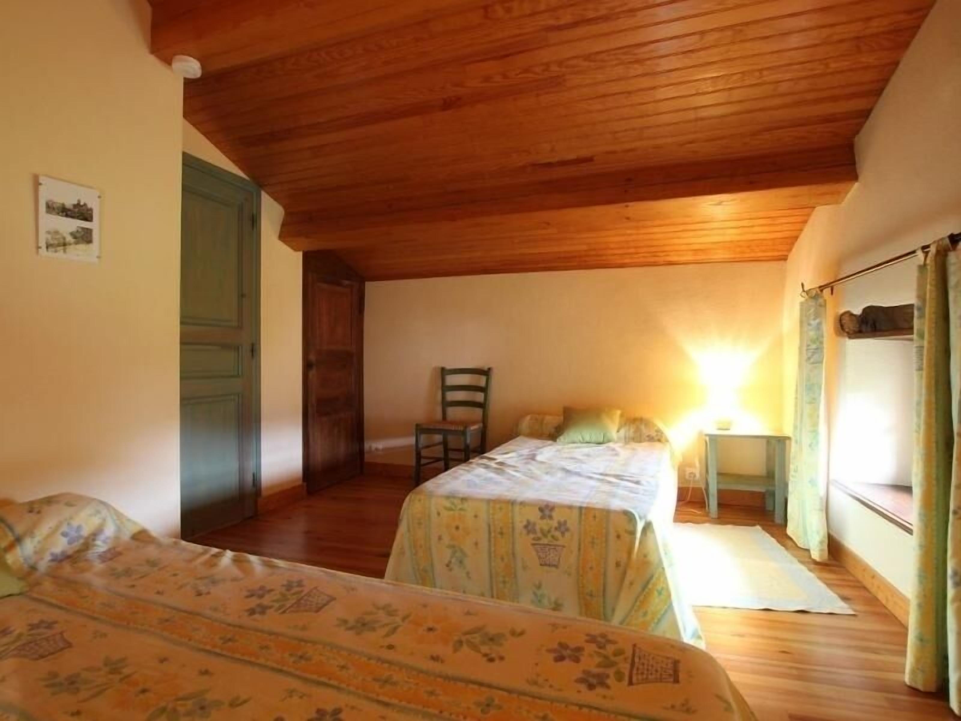 A room with two beds, wooden ceiling, and a small table with a lamp.