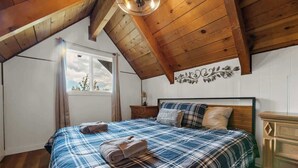 Une chambre avec un plafond en bois, un lit recouvert d’une couverture à carreaux, une table de chevet et une commode.