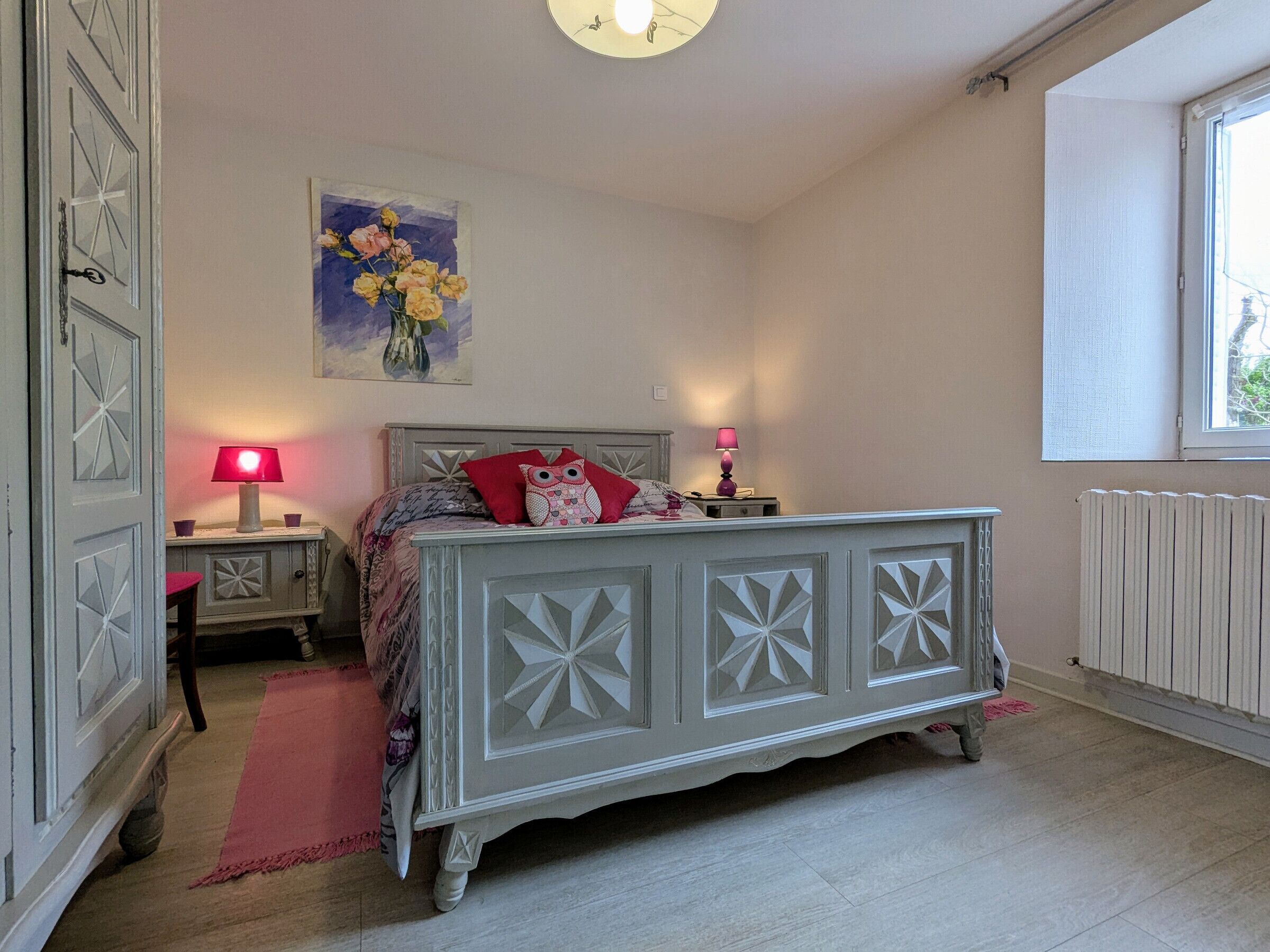 A bedroom with a bed, a nightstand, a lamp, a painting, and a window.