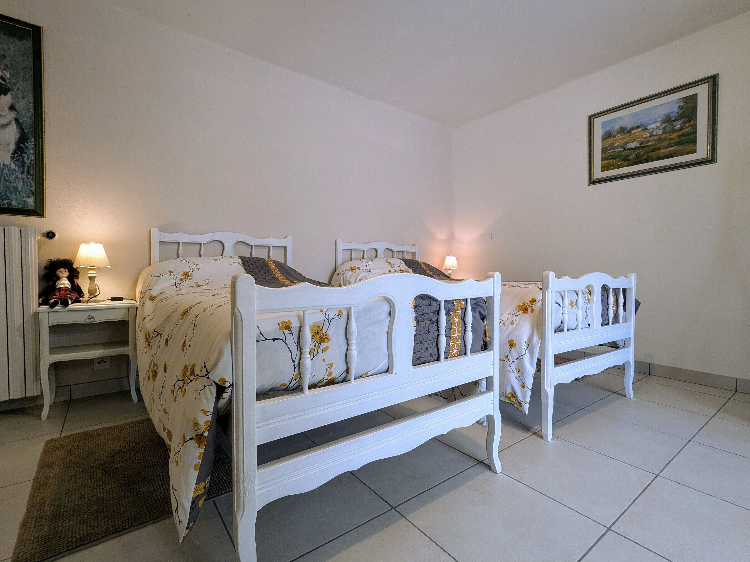 A bedroom with two white wooden beds, a nightstand with a lamp, and a framed painting on the wall.