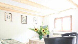 Uma sala de estar com sofá, mesa de centro e televisão. Há quadros nas paredes e uma janela com persianas.