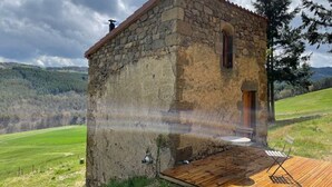 Edificio rústico de piedra con terraza de madera y una sola puerta, rodeado de un paisaje cubierto de césped y colinas.