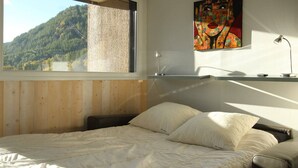 A bedroom with a large window, a bed, a desk, and a painting.