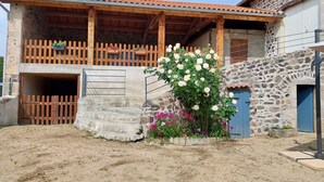 A rustic stone building with a wooden balcony, a blue door, and a garden with white and pink flowers.