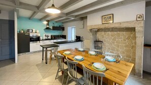 A dining area with a wooden table set for four, a stone fireplace, and a kitchen in the background.