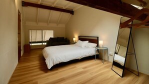 Une chambre avec un plancher en bois, un lit avec une literie blanche, une table de chevet avec une lampe, un grand miroir et une fenêtre avec des stores.