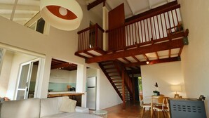 Un vaste espace de vie doté d’un haut plafond, d’un escalier en bois, d’un canapé, d’une table à manger avec des chaises et, au fond, d’une petite cuisine.
