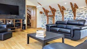 A modern living room with a black leather sofa, a coffee table, and a TV mounted on the wall. There is a wooden entertainment unit with electronic devices and a stone wall with wooden beams. - Luxury Gîte with Spa, Terrace, and Game Room in Scenic Blanlhac, Rosières (Rosières)