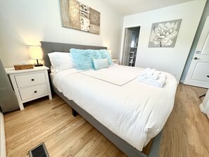 Een opgemaakt bed met witte lakens en blauwe kussens, een nachtkastje met een lamp, en een muur met twee schilderijen.