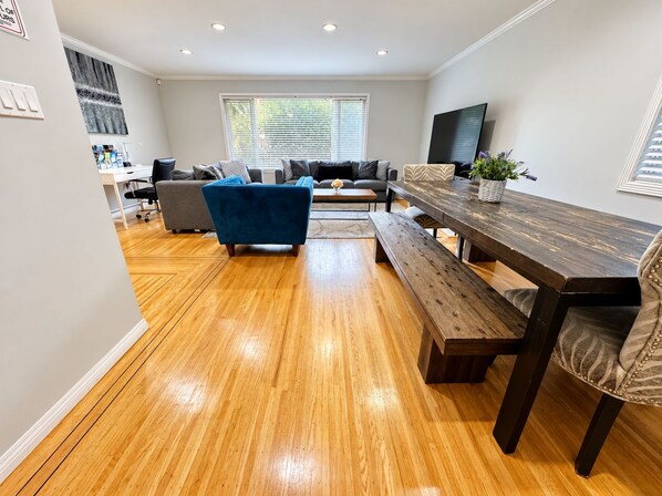 Un salón moderno con suelo de madera, un sofá azul, una mesa de comedor de madera con bancos y un ventanal con persianas.