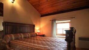 Une chambre avec un plafond en bois, un lit recouvert d’une couette à rayures, une table de chevet avec une lampe et une fenêtre avec des rideaux.