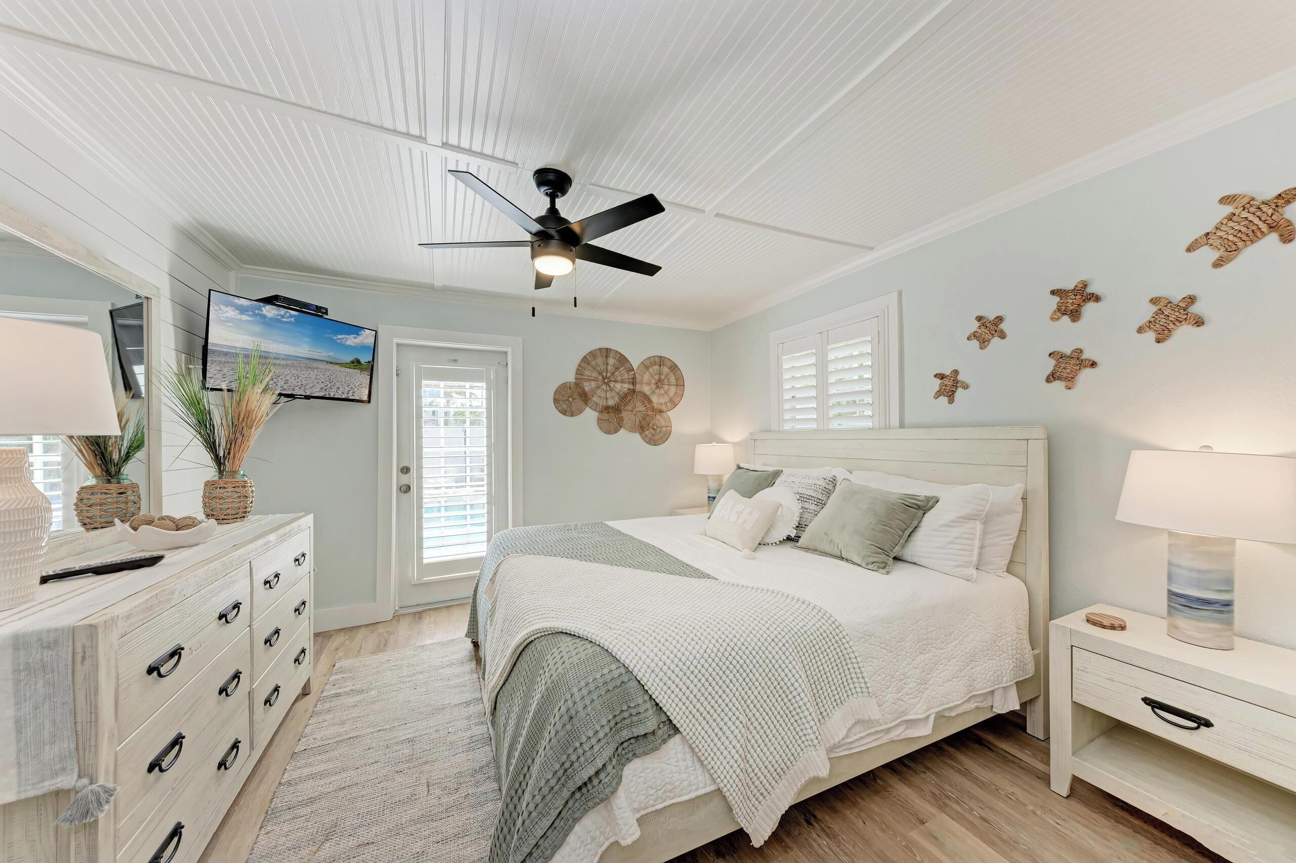 Un dormitorio con cama, mesita de noche, cómoda, ventilador de techo, televisor y decoración en la pared.