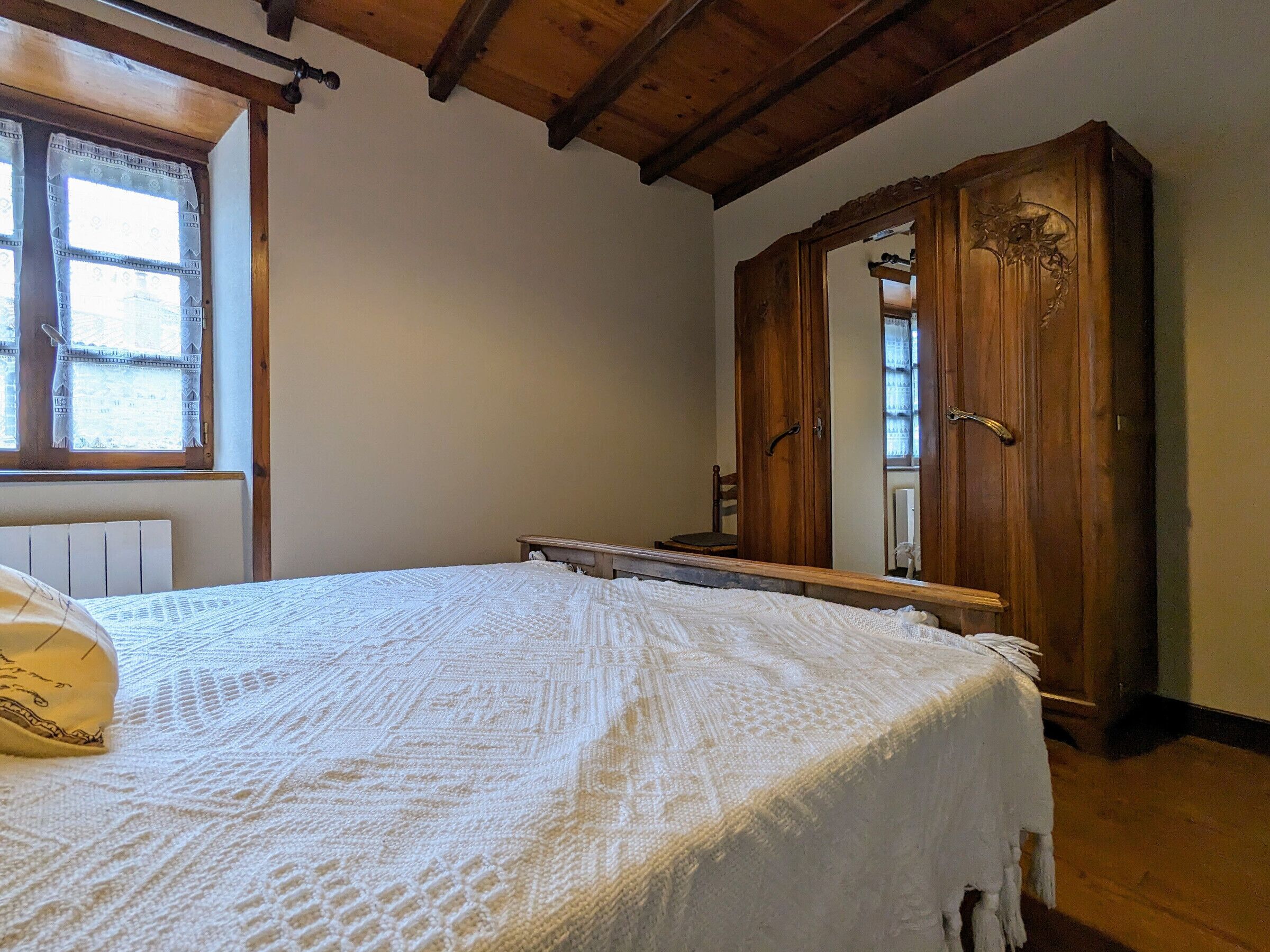 Un dormitorio con techo de madera, una cama con manta blanca, un armario de madera y un ventanal con cortinas estampadas.
