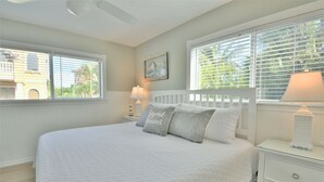 A bedroom with a large bed, white bedding, a nightstand with a lamp, and a window with blinds.