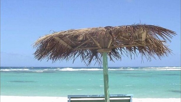 A beach umbrella with a wooden frame and a thatched top, a wooden bench, and a clear view of the ocean.