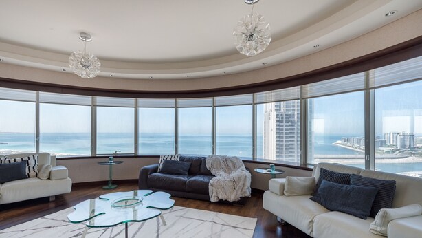 Ein geräumiges Wohnzimmer mit gewölbter Decke, großen Fenstern mit Blick auf das Meer und die Stadt sowie modernen Möbeln, darunter ein weißes Sofa, ein dunkles Sofa und ein Glastisch.