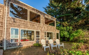 Une maison en bois à deux étages, dotée de grandes fenêtres et d’une terrasse aménagée avec des fauteuils Adirondack.