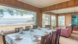 A dining area with a table set for four, large windows offering a view of the beach, wooden paneled walls, and a pink sofa in the background. - Oceanfront home in Newport with sweeping views, and easy beach access (Newport)