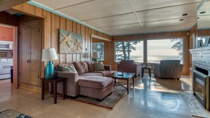 Un salon spacieux avec un grand canapé d’angle, une table basse et une cheminée. La pièce est dotée de lambris en bois et de grandes fenêtres offrant une vue sur l’océan.