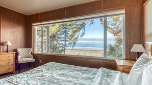 Une chambre avec une grande fenêtre donnant sur une plage, un lit recouvert d’une couvre-lit à motifs, une chaise et une table de chevet avec une lampe.