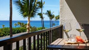 Un balcon avec vue sur l’océan et les palmiers, deux verres à vin et une bougie posés sur une table en bois.