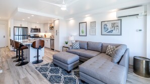 Un salon moderne avec un canapé d’angle gris, un coin bar avec des tabourets, une cuisine avec des meubles blancs et un ventilateur de plafond.