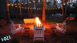 Un fuoco da campo con due poltrone Adirondack rivolte verso di esso, circondato da alberi e luci decorative.