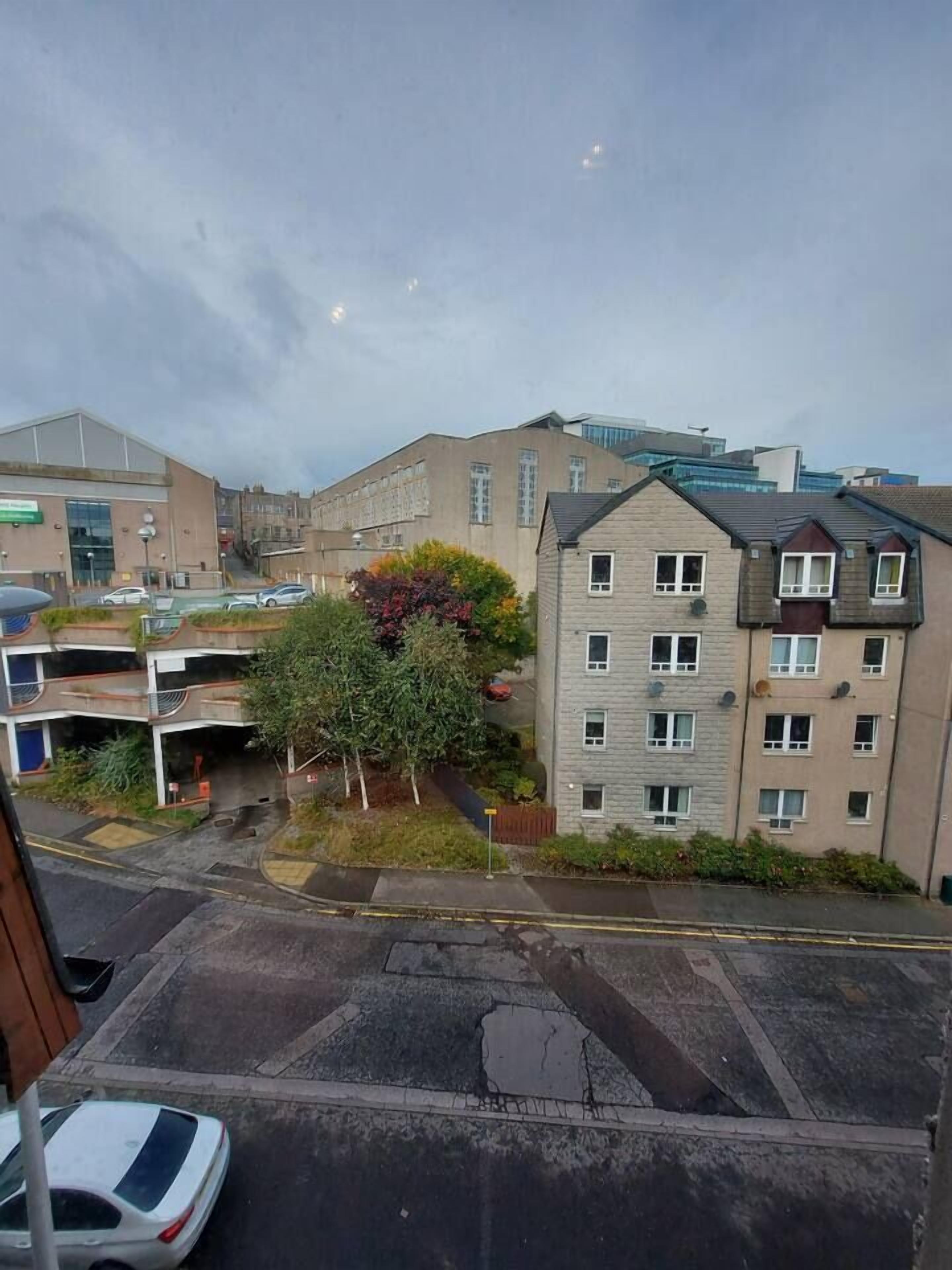 Um edifício residencial de vários andares com telhado plano, área de estacionamento e uma árvore em primeiro plano.