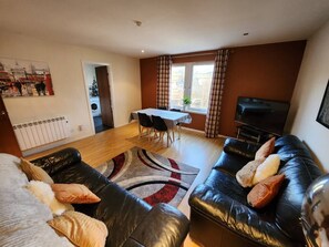 Ein Wohnzimmer mit schwarzem Ledersofa, einem Esstisch mit Stühlen, einem Fernseher und einer Heizung.