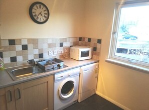 Compact kitchen with microwave, stainless steel sink, and washing machine, offering a view of the outside.