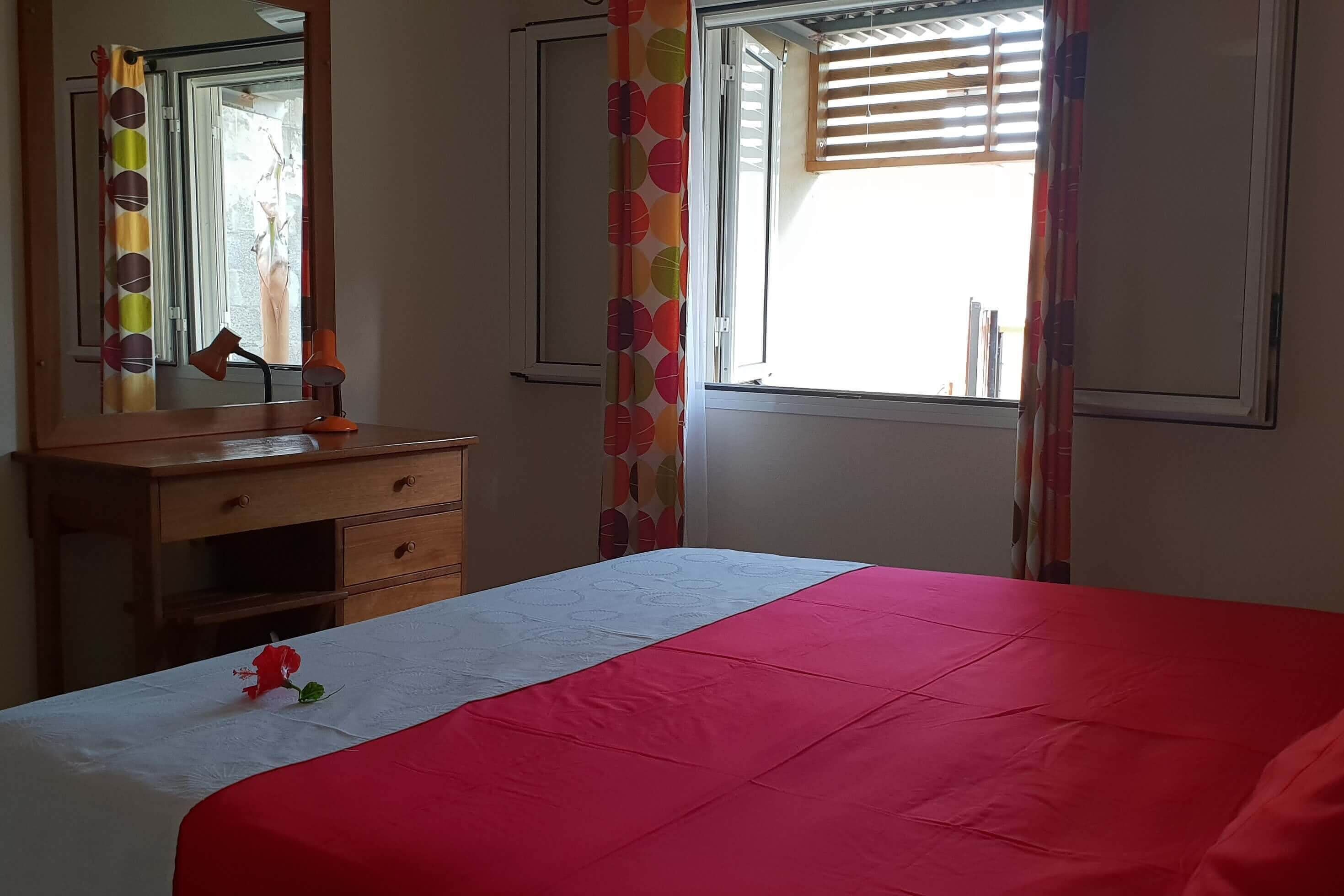 A bedroom with a bed, a nightstand, a mirror, a window with curtains, and a view of the outside.