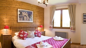 Een slaapkamer met houten lambrisering, een bed, twee nachtkastjes, een rode lamp, een raam met gordijnen en een schilderij van een berg aan de muur.
