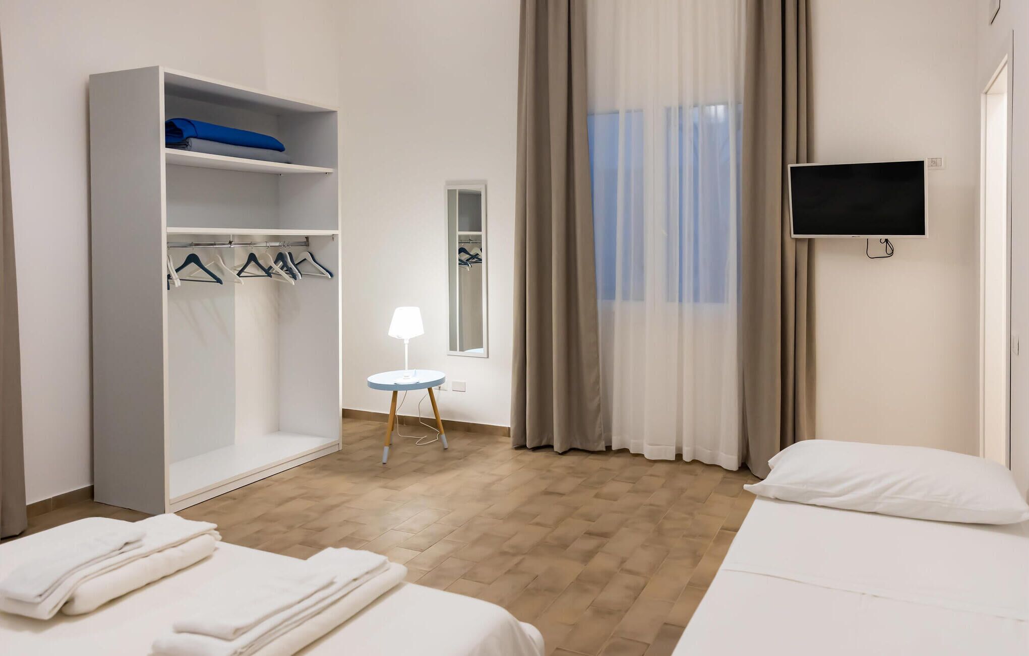 Ein Hotelzimmer mit einem Bett, einem Kleiderschrank, einem kleinen Tisch mit Lampe, einem Fernseher und einem Fenster mit Vorhängen.