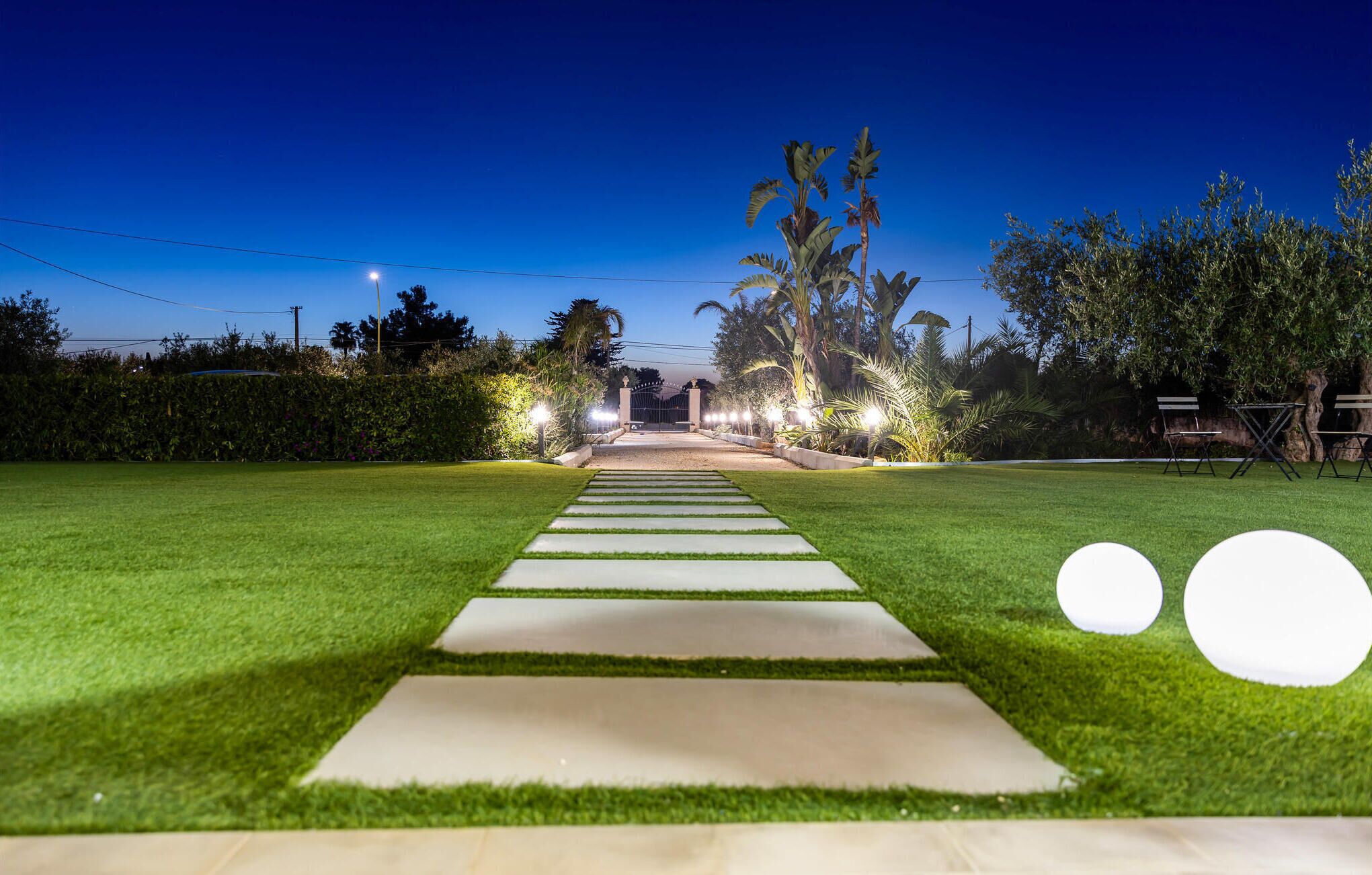 A well-lit pathway leading to a gated entrance surrounded by lush greenery and palm trees.