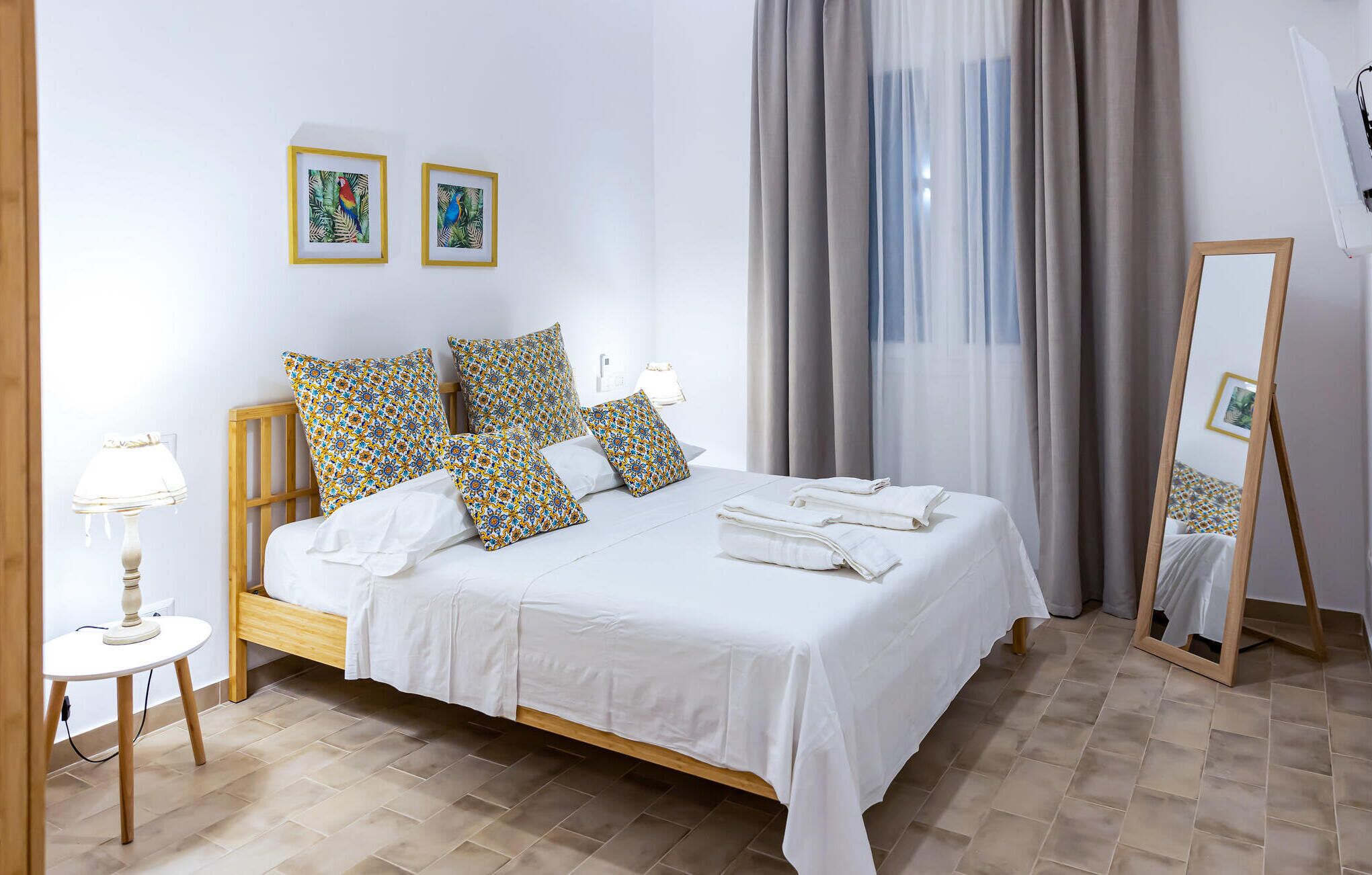 A neatly made bed with white linens, a wooden headboard, and patterned pillows. There are two framed pictures on the wall above the bed, a lamp on a bedside table, and a mirror leaning against the wall.