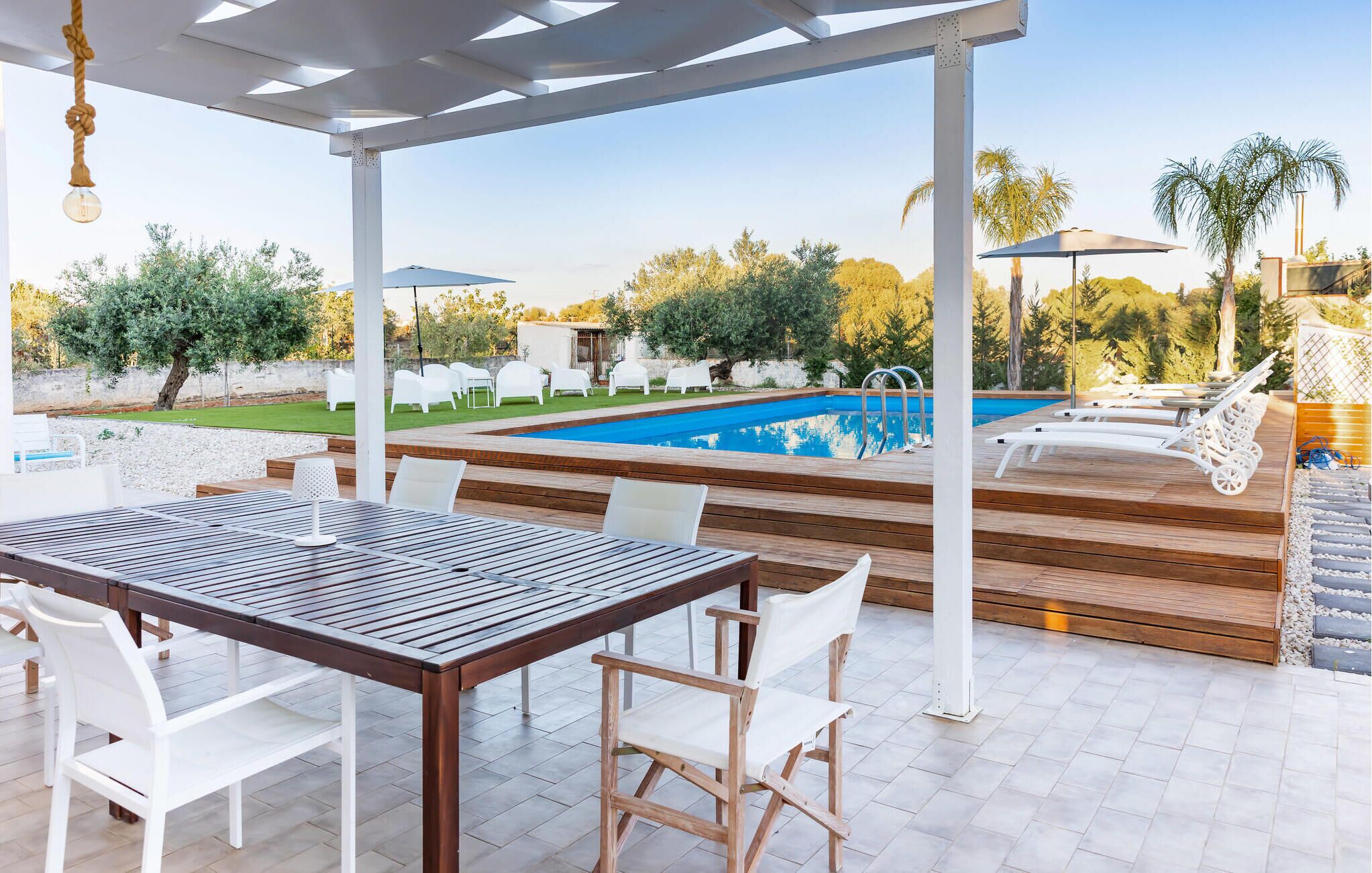 An outdoor patio with a pool, wooden deck, white chairs, and a pergola.