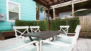 Une terrasse avec une table et des chaises blanches, une maison bleue en arrière-plan et une clôture en bois.