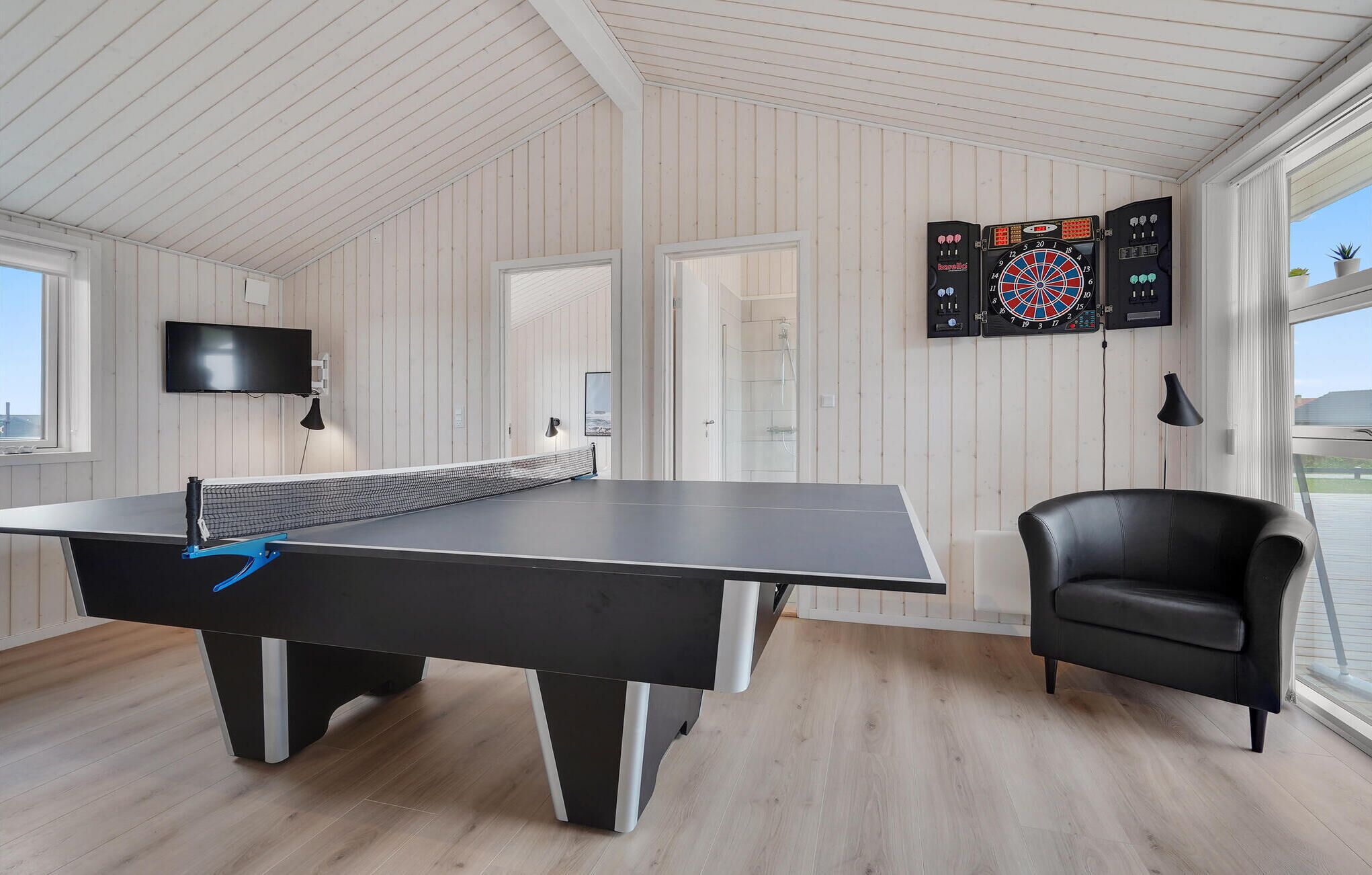 Una mesa de ping pong, un dardo, un televisor y una silla en una habitación con pisos de madera y paredes blancas.
