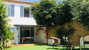 Un patio con césped artificial, mobiliario de mimbre y un edificio con contraventanas azules.