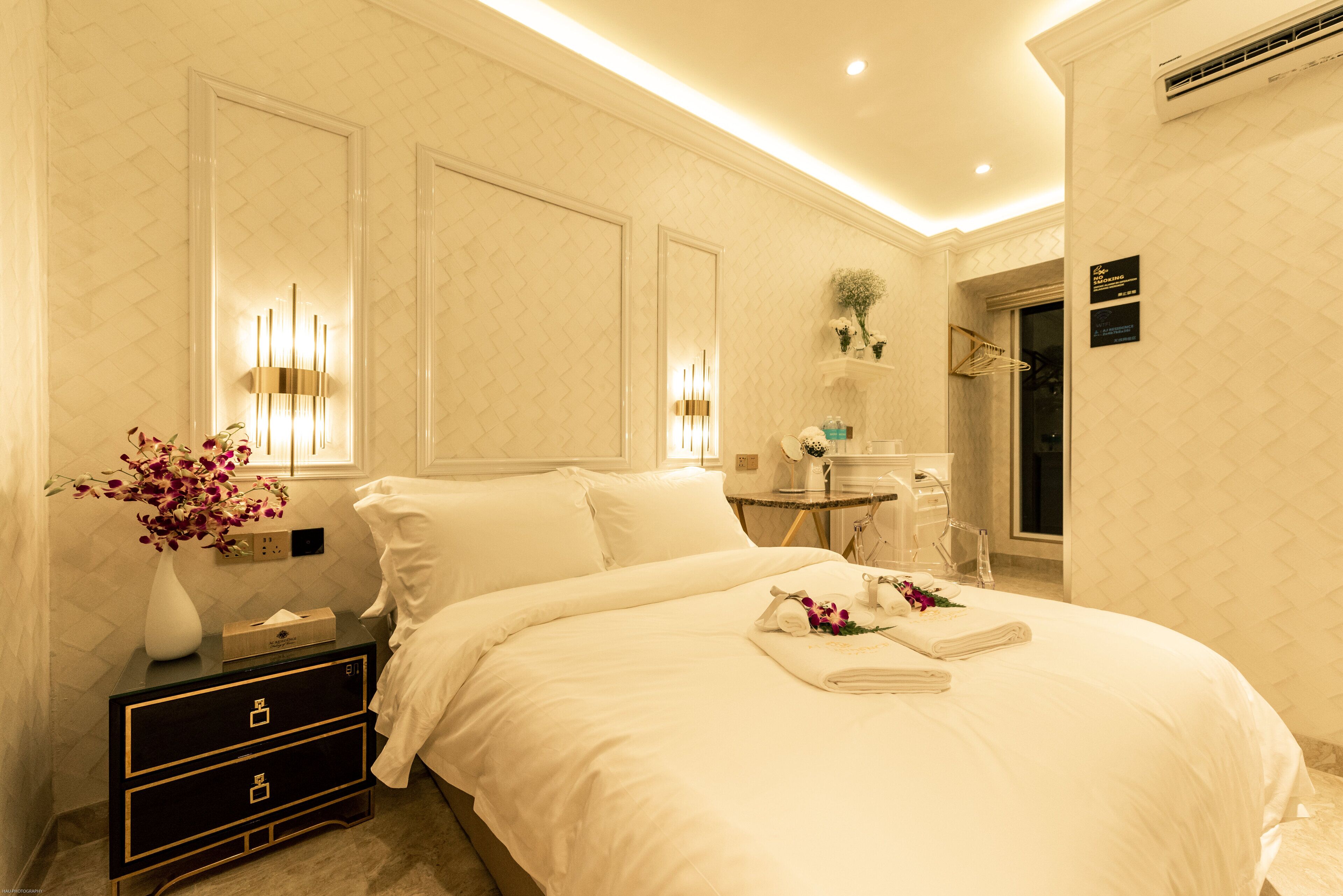 A hotel room with a neatly made bed, a bedside table with a vase of flowers, and a wall-mounted mirror.