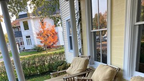 A covered porch with wicker furniture and cushions, overlooking a residential area with a tree and a building in the background.