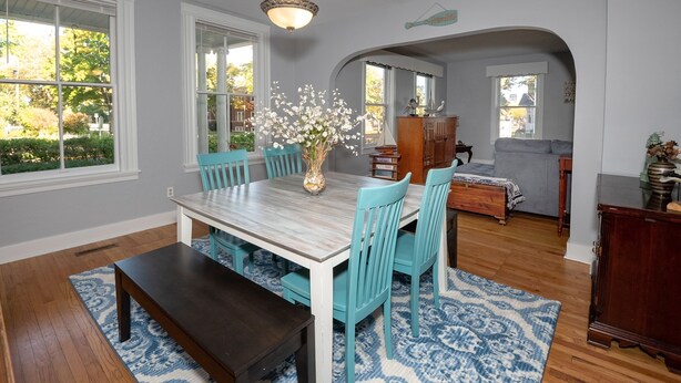A dining room with a wooden table, blue chairs, and a vase of flowers. There is a bench, a rug, and a fireplace in the background.