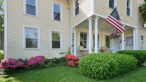 A two-story house with a porch, a hanging American flag, and a well-maintained garden with pink flowers and green bushes.