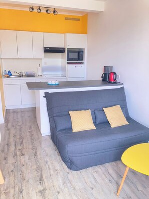 A compact living space with a kitchenette, a grey sofa with two cushions, and a yellow table.