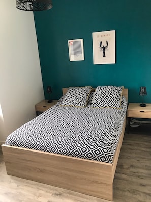 A bedroom with a bed, two nightstands, a wall-mounted shelf, and two framed pictures.
